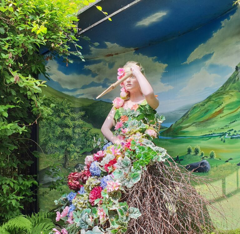 a statue woman with blonde hair and a green dress with colourful flowers which tells one of the Lost Legends of the Stone Circle at King Arthur's Labyrinth in Corris